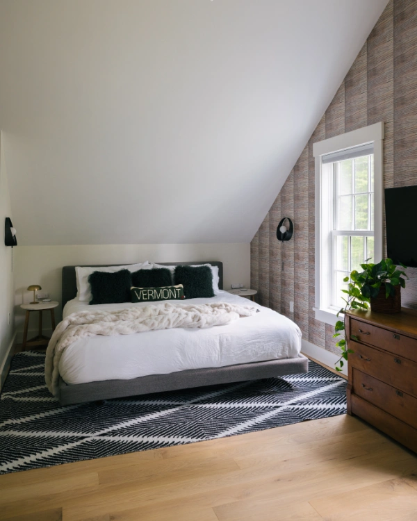 Angled bedroom view highlighting vaulted ceiling, neutral bedding, Vermont-themed accent pillow, and large window framing greenery